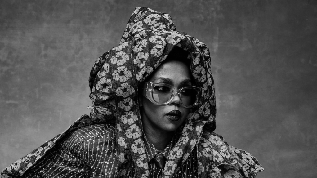 A person wearing a hooded floral jacket and glasses, with a serious expression, against a plain dark background.
