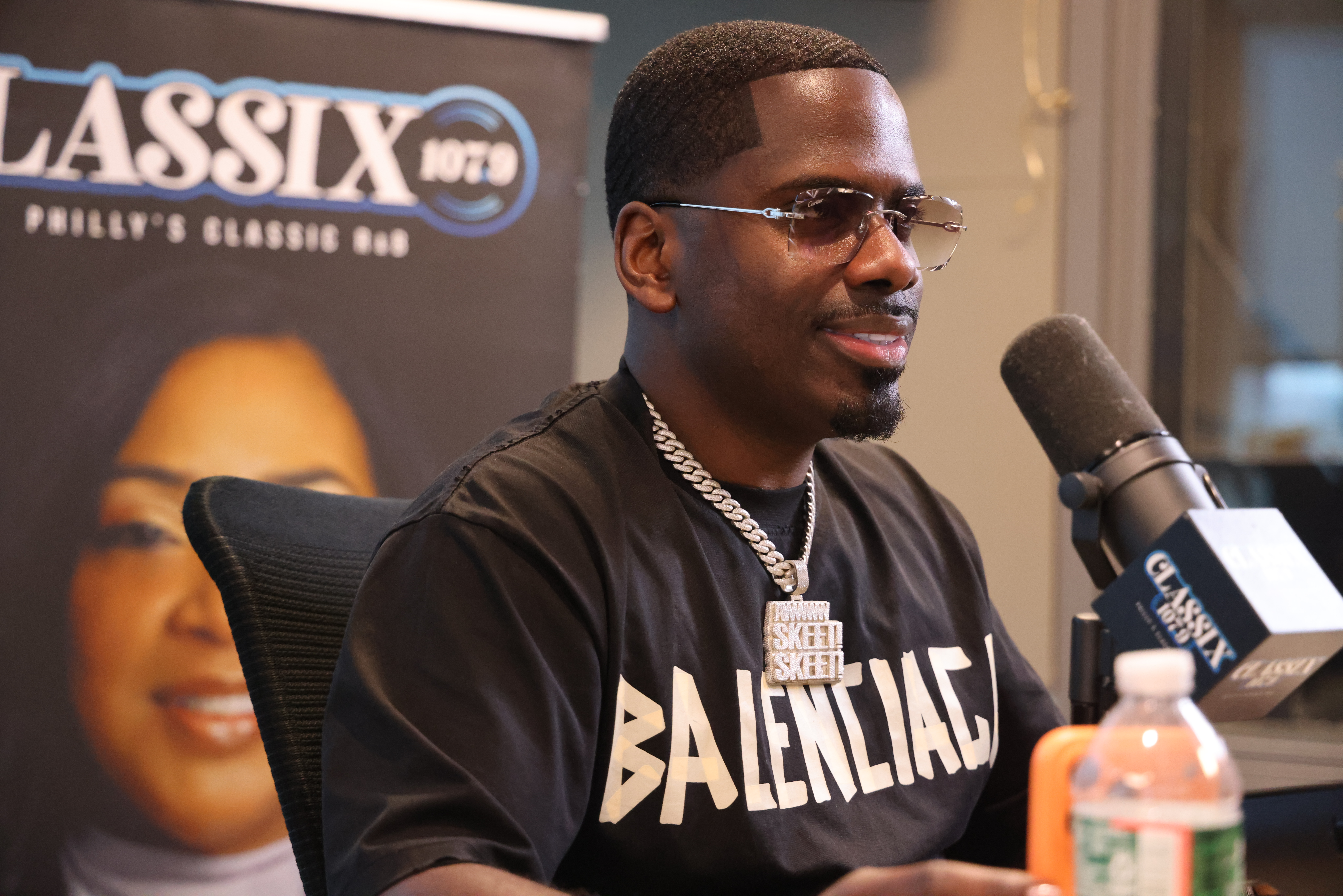 A man wearing a black "Balentiac" t-shirt and sunglasses is being interviewed in front of a "Classix 100.3" logo backdrop.
