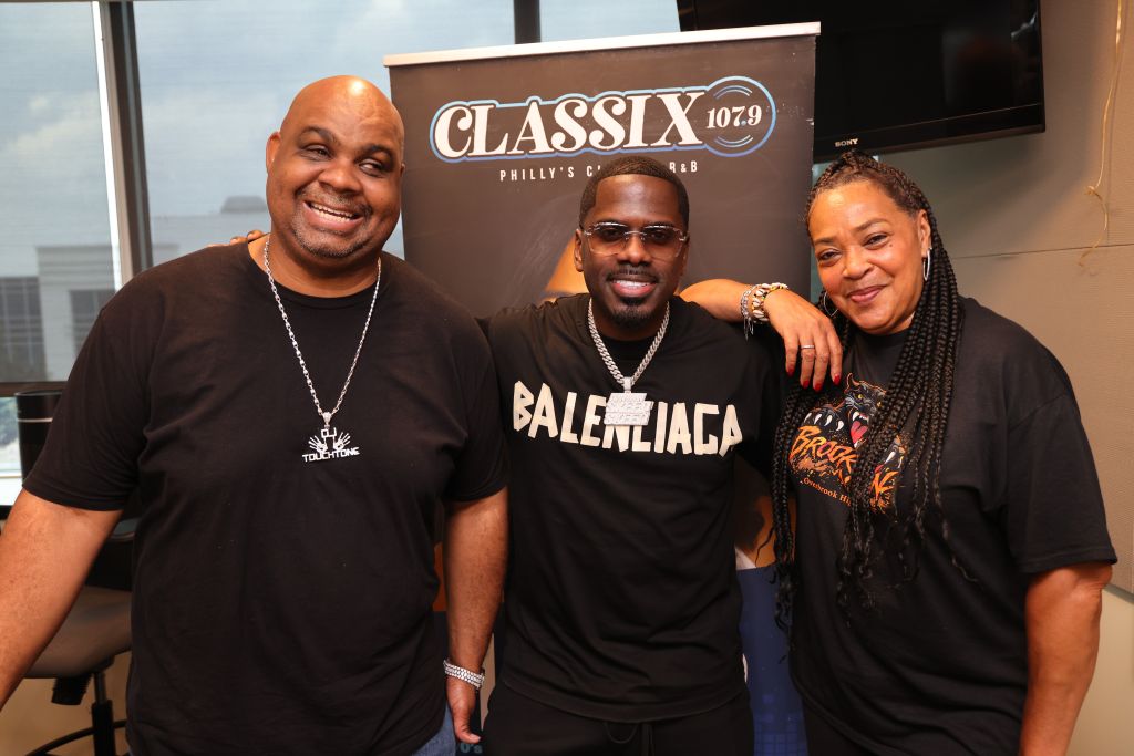 Three people posing in front of a Classix 107.9 radio station banner, one wearing a Balenciaga t-shirt.