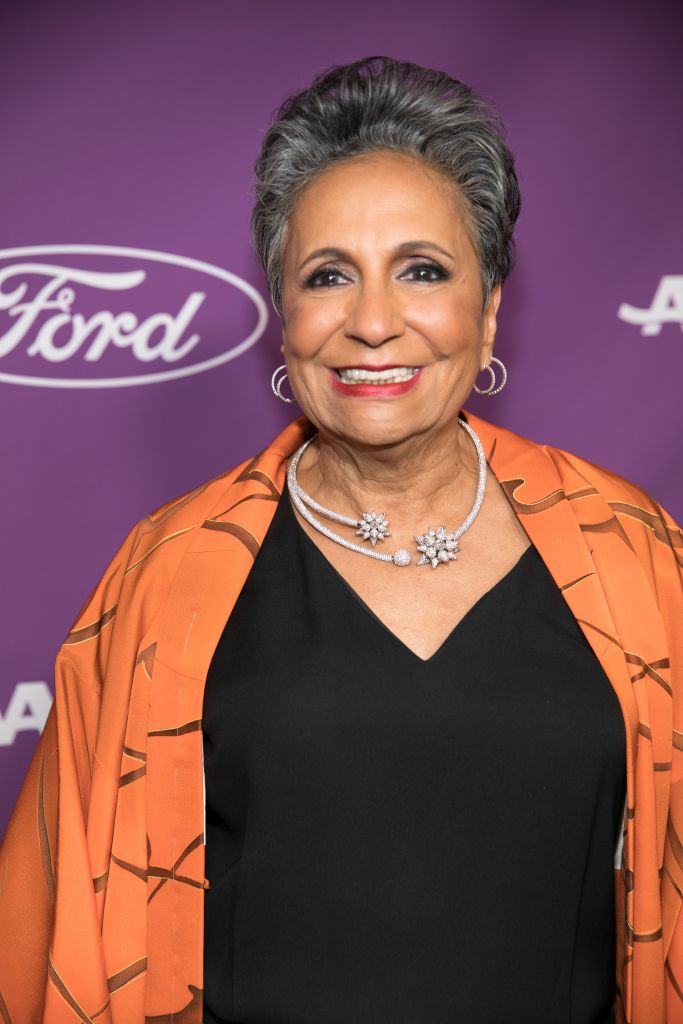 A smiling older woman wearing an orange patterned jacket and black dress, with diamond jewelry, standing in front of a purple background with the Ford logo.