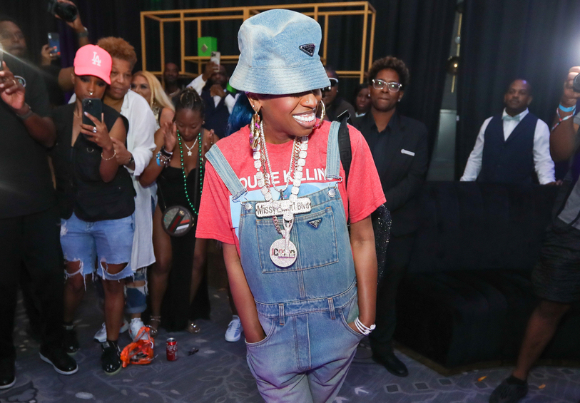 A person wearing a pink bucket hat, red t-shirt, and denim overalls standing in a crowd of people at what appears to be an event or concert.