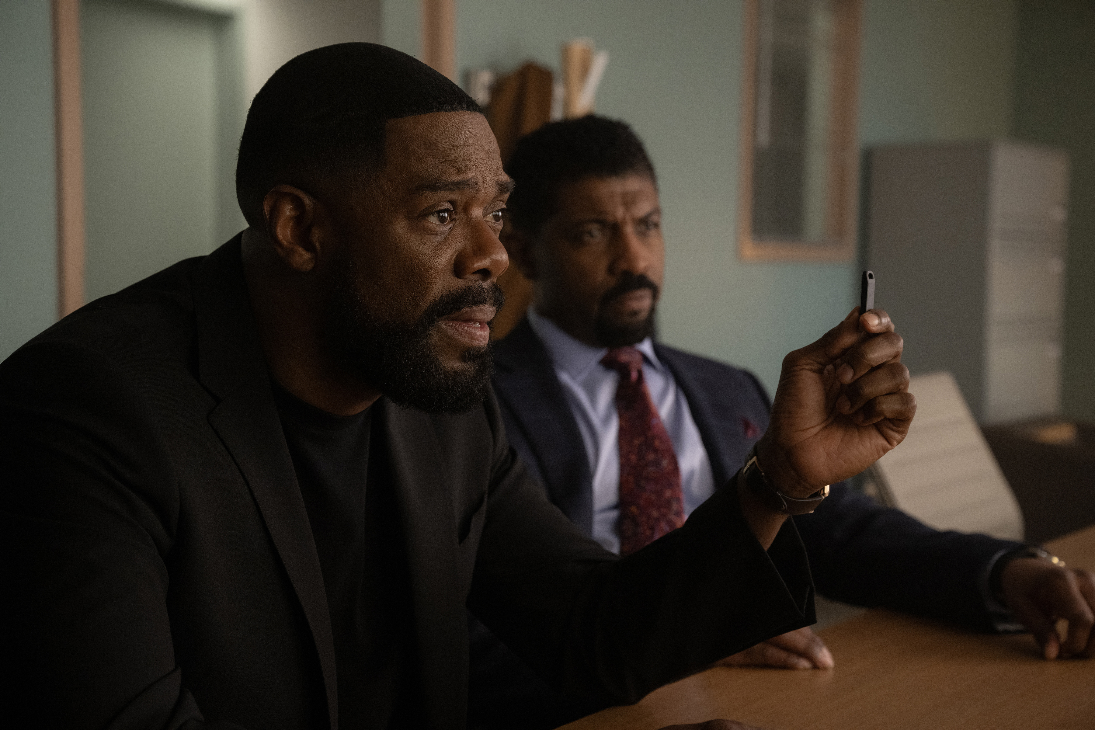 Two serious-looking Black men in suits, one holding a smartphone, in a dimly lit room.