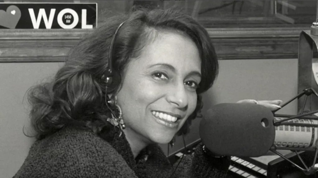 A smiling woman wearing headphones and sitting in a radio studio, with the call letters "WOL" visible on the wall behind her.