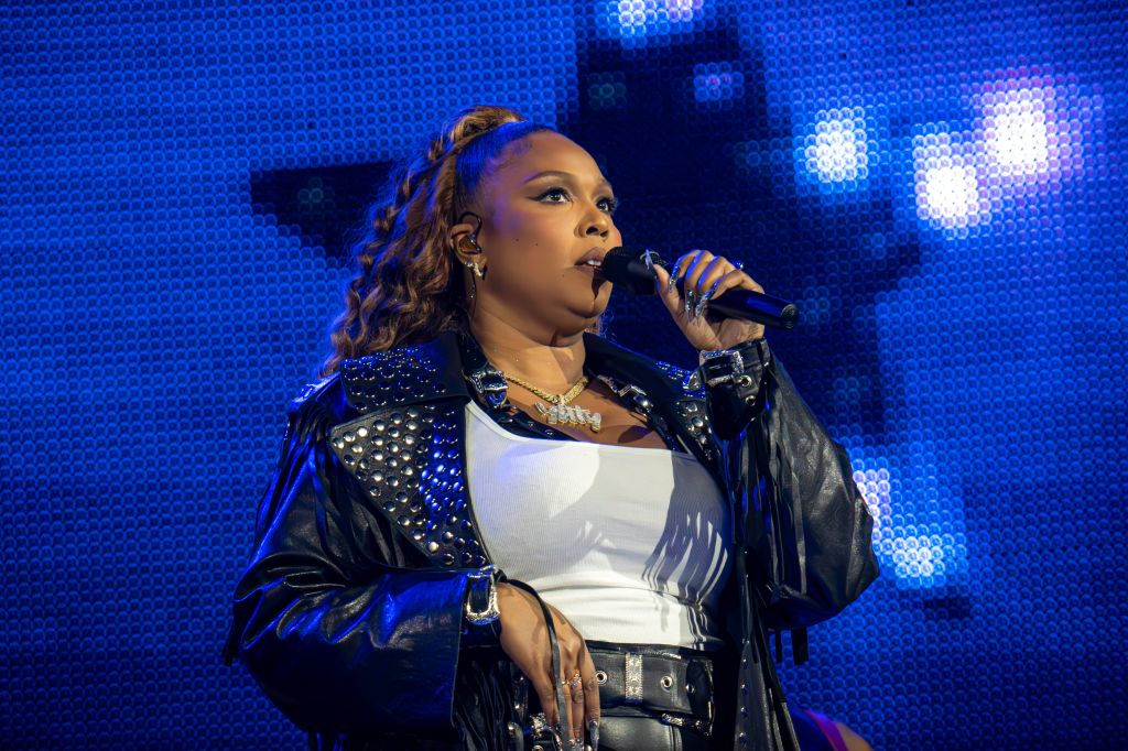 A woman with curly hair wearing a studded black jacket singing into a microphone on a brightly lit stage.