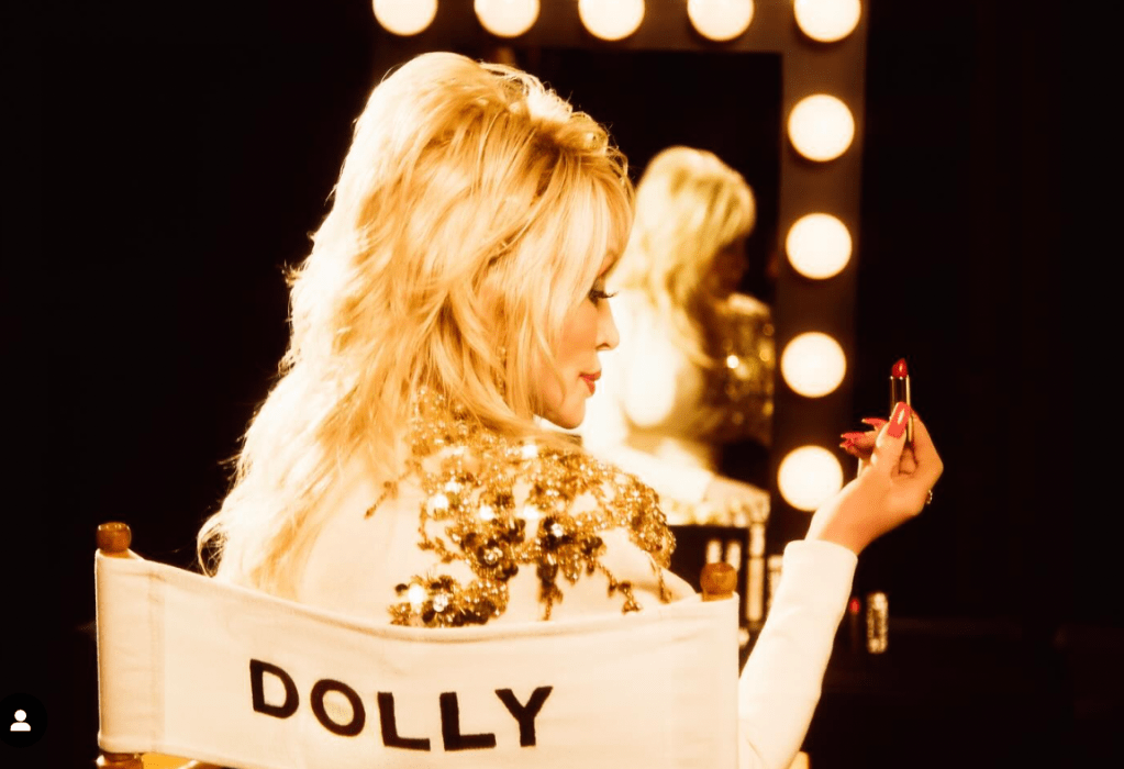 A woman with long blonde hair applying lipstick in front of a mirror with lights around it, wearing a gold sequined jacket.