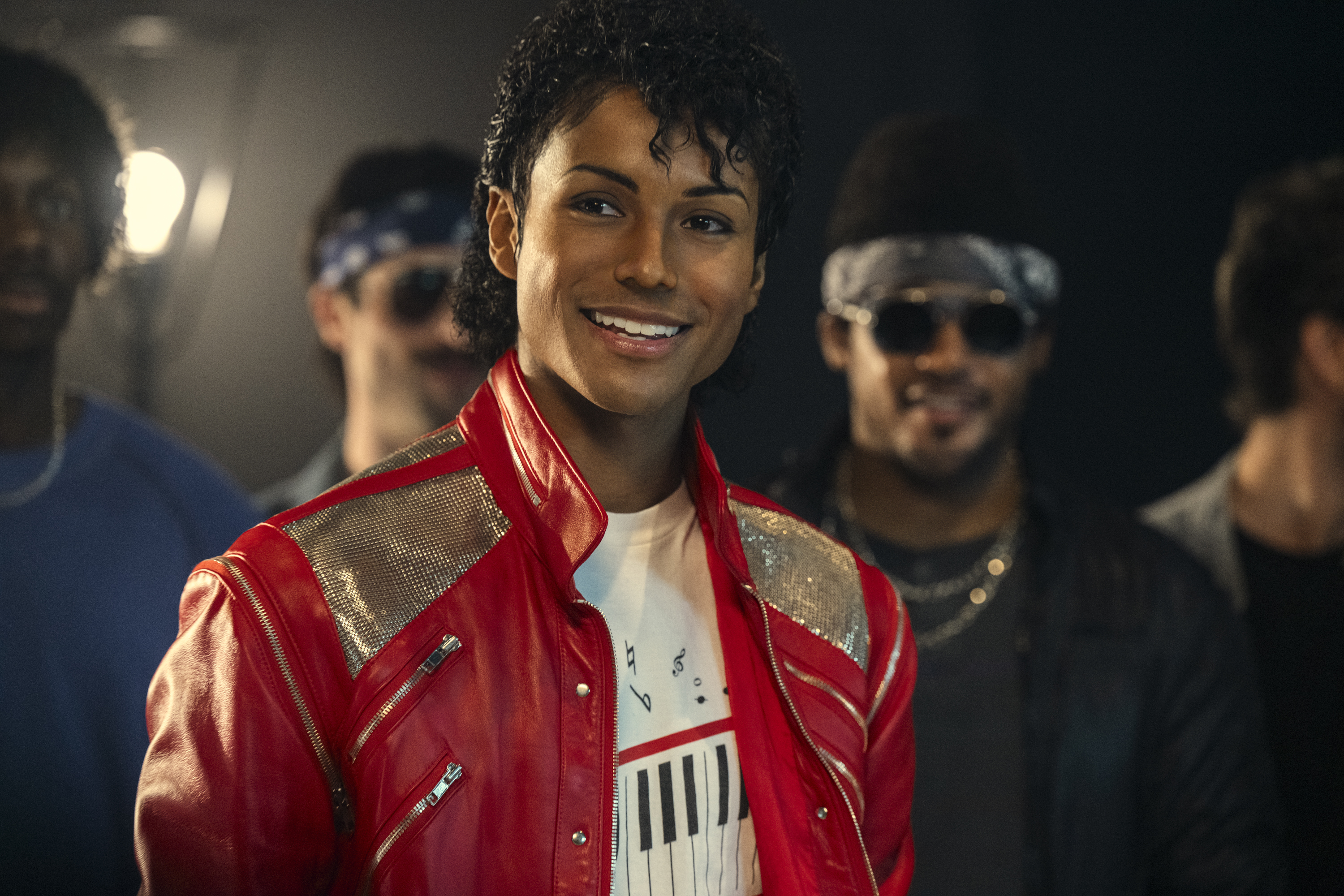 A smiling man in a red leather jacket with sequins, standing in front of other people wearing sunglasses.