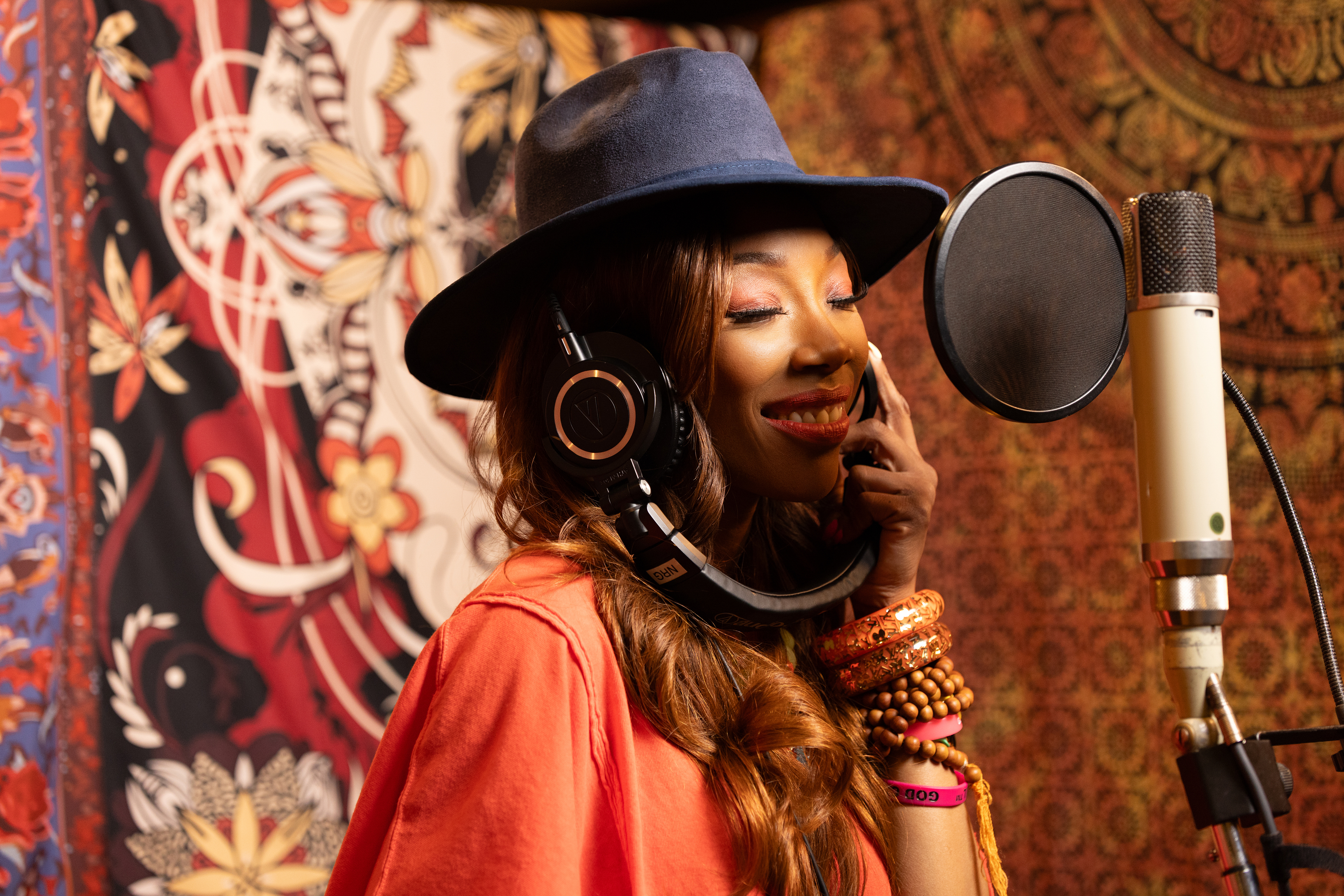 "Woman wearing a hat and headphones singing into a studio microphone, smiling, with colorful patterned tapestries in the background, suggesting a creative, artistic music recording environment."