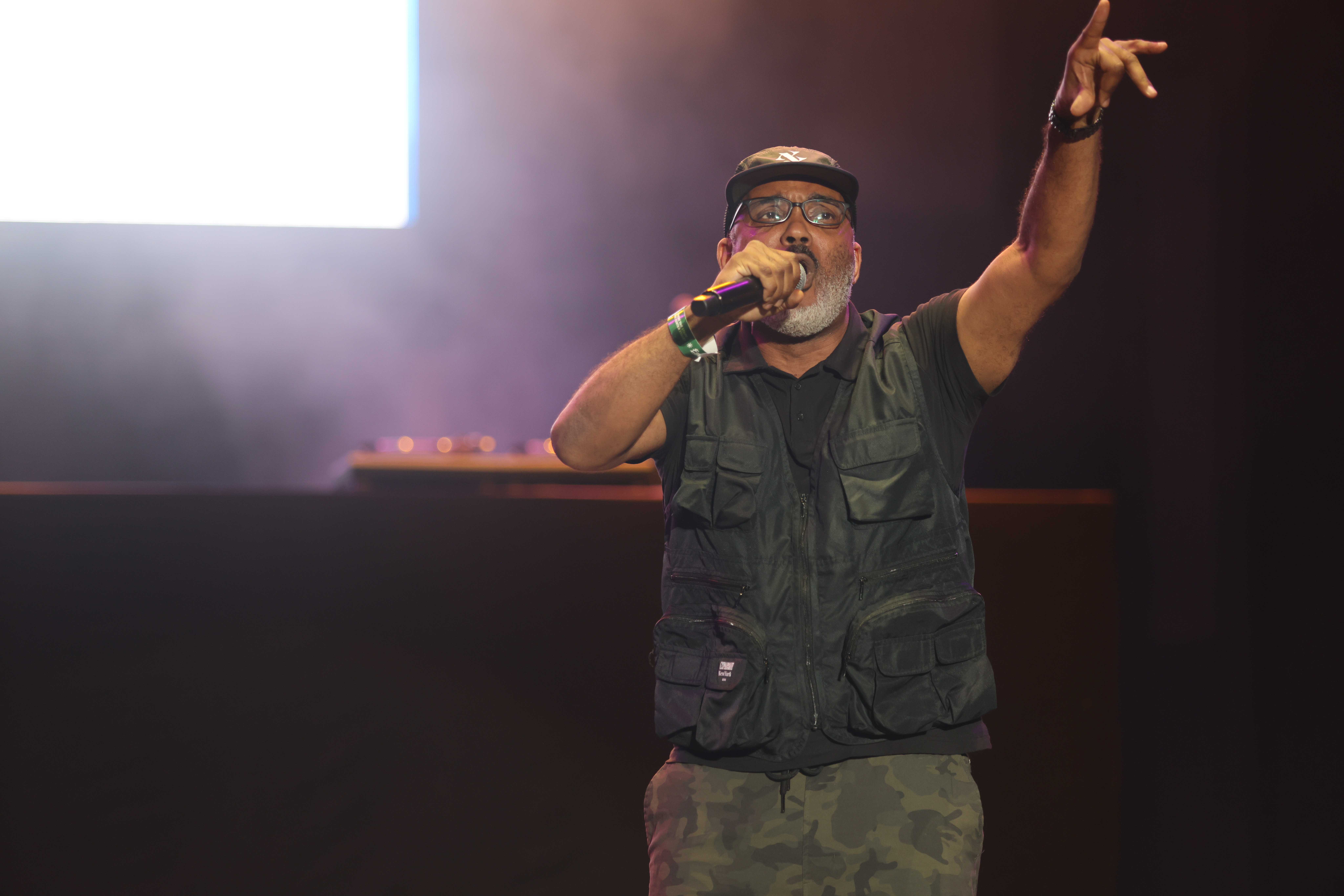 A man wearing a black vest and hat performing on stage, holding a microphone and gesturing with his hand.