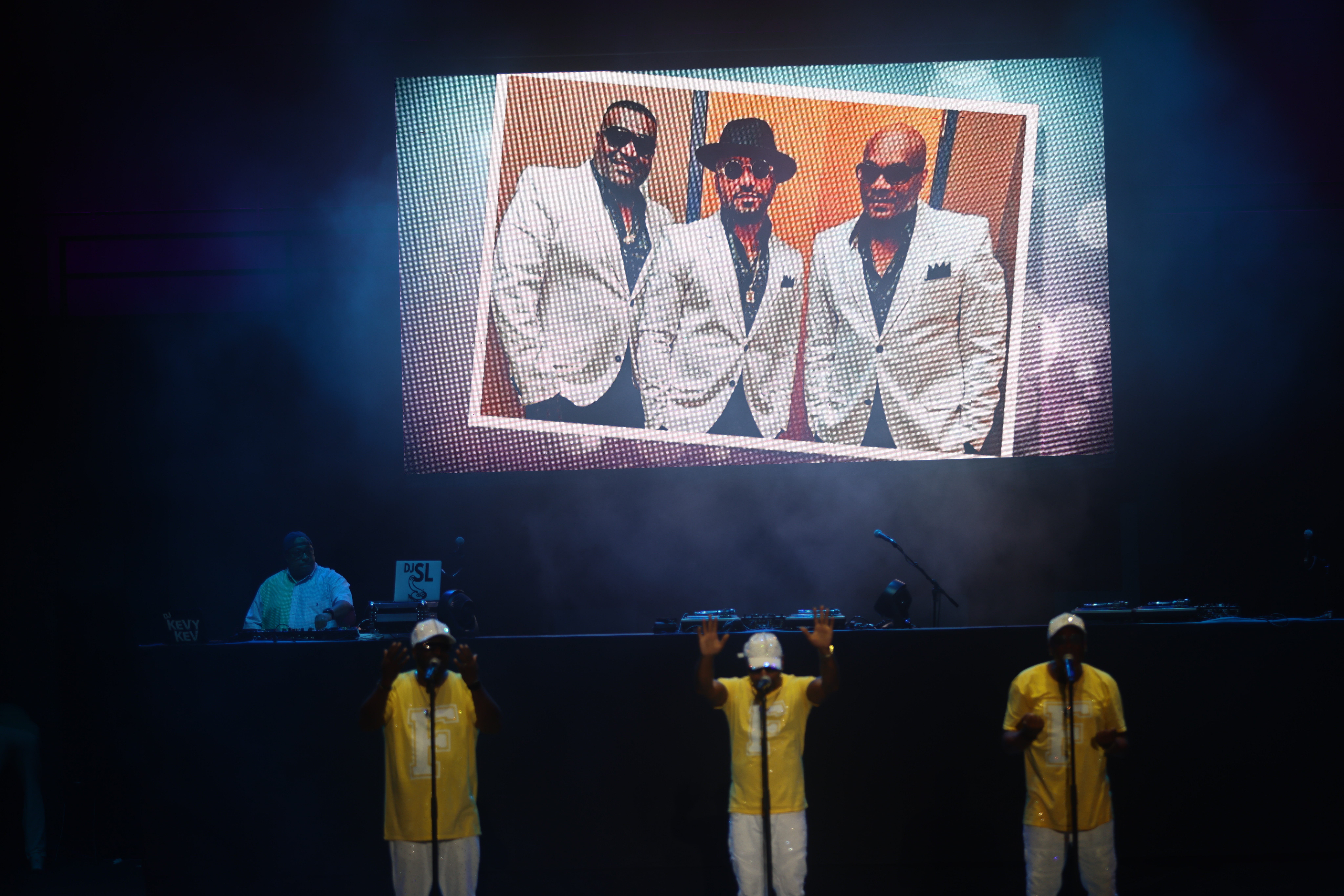 Three men in white suits standing together on a stage, with a large projection screen behind them displaying their image.