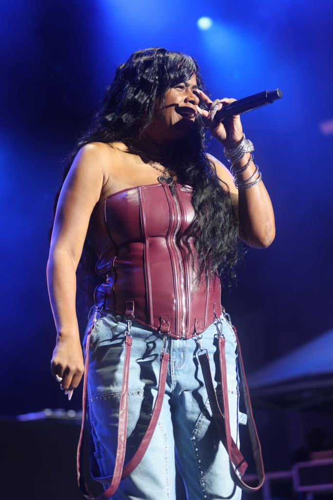 A woman with long dark hair singing into a microphone on stage, wearing a burgundy leather corset-style top and jeans with straps.