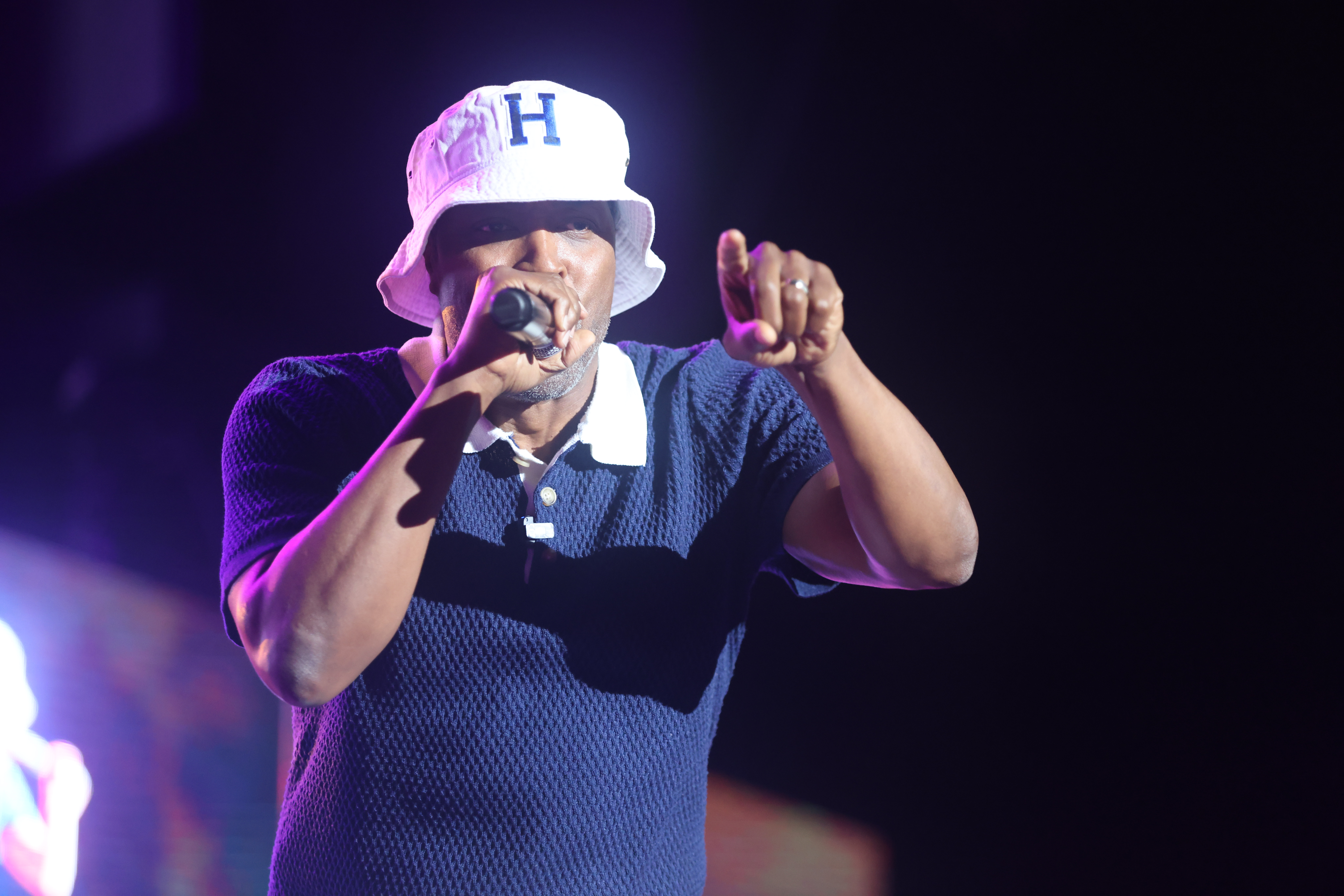 A person wearing a white hat with the letter "H" performing on stage in a dark setting, holding a microphone.