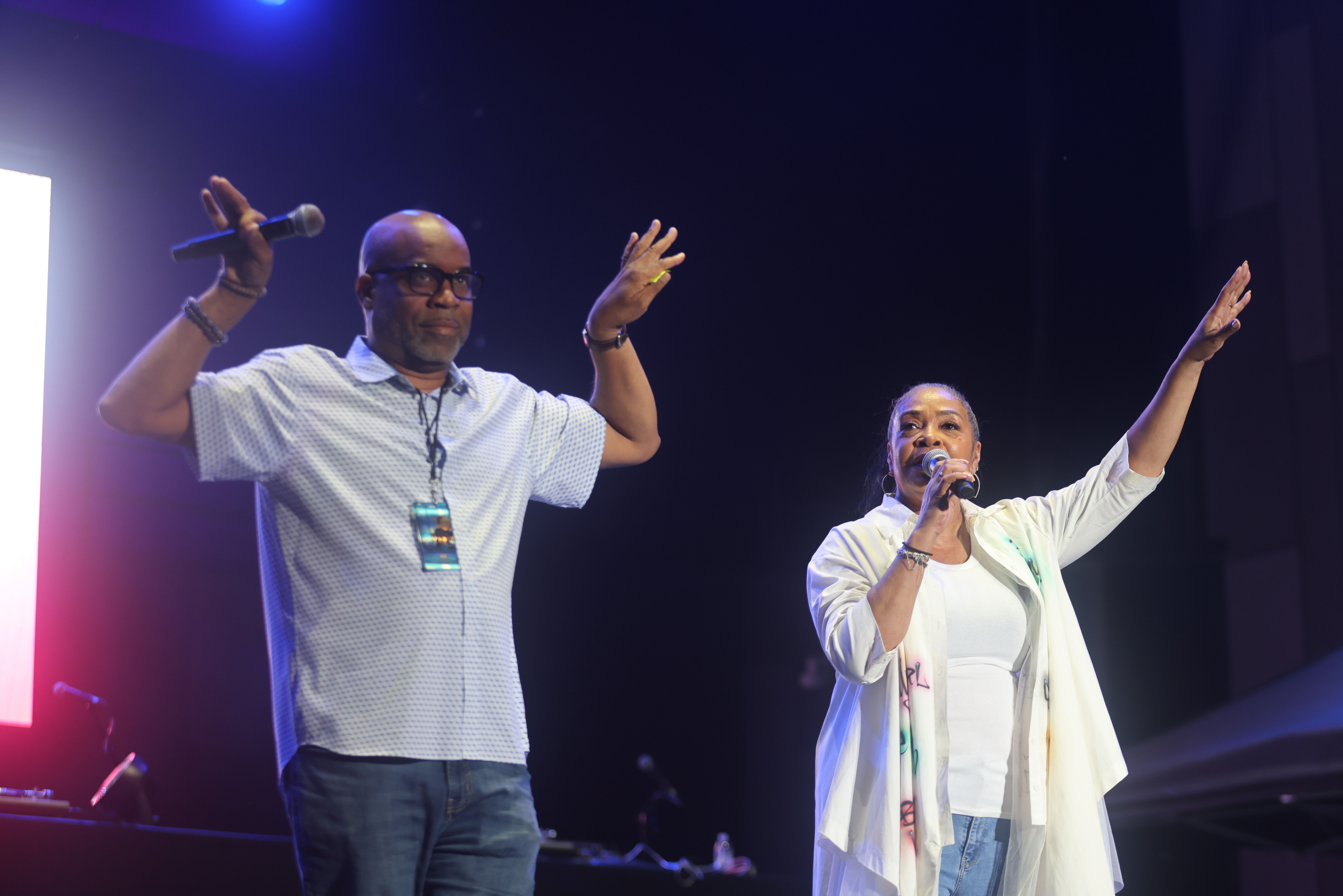 Two people performing on stage, a man with his arms raised and a woman singing into a microphone.