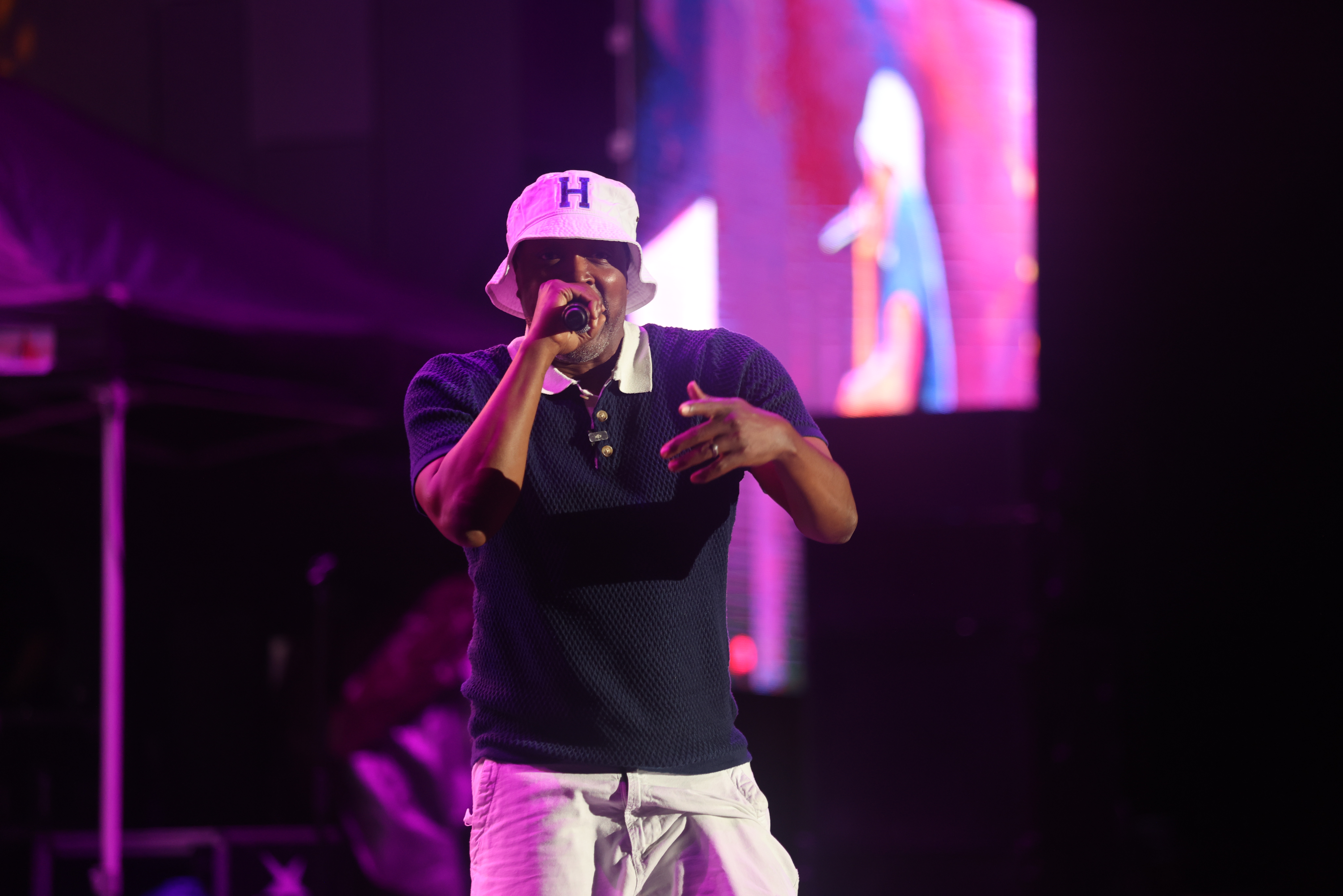 A performer wearing a pink "H" cap singing into a microphone on a brightly lit stage with colorful lighting effects.