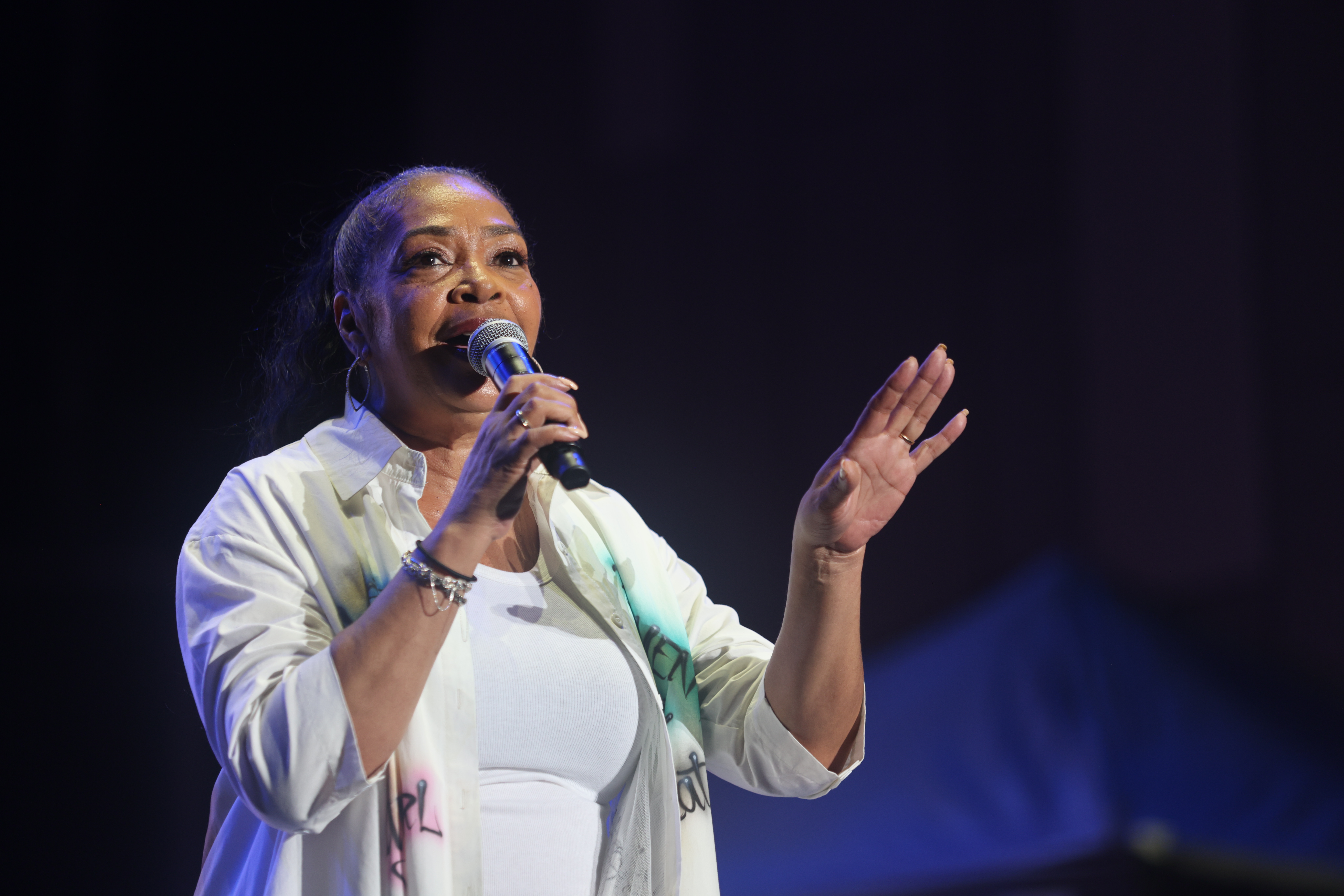 A woman with long dark hair singing into a microphone on a dimly lit stage, wearing a white jacket and gesturing with her hand.