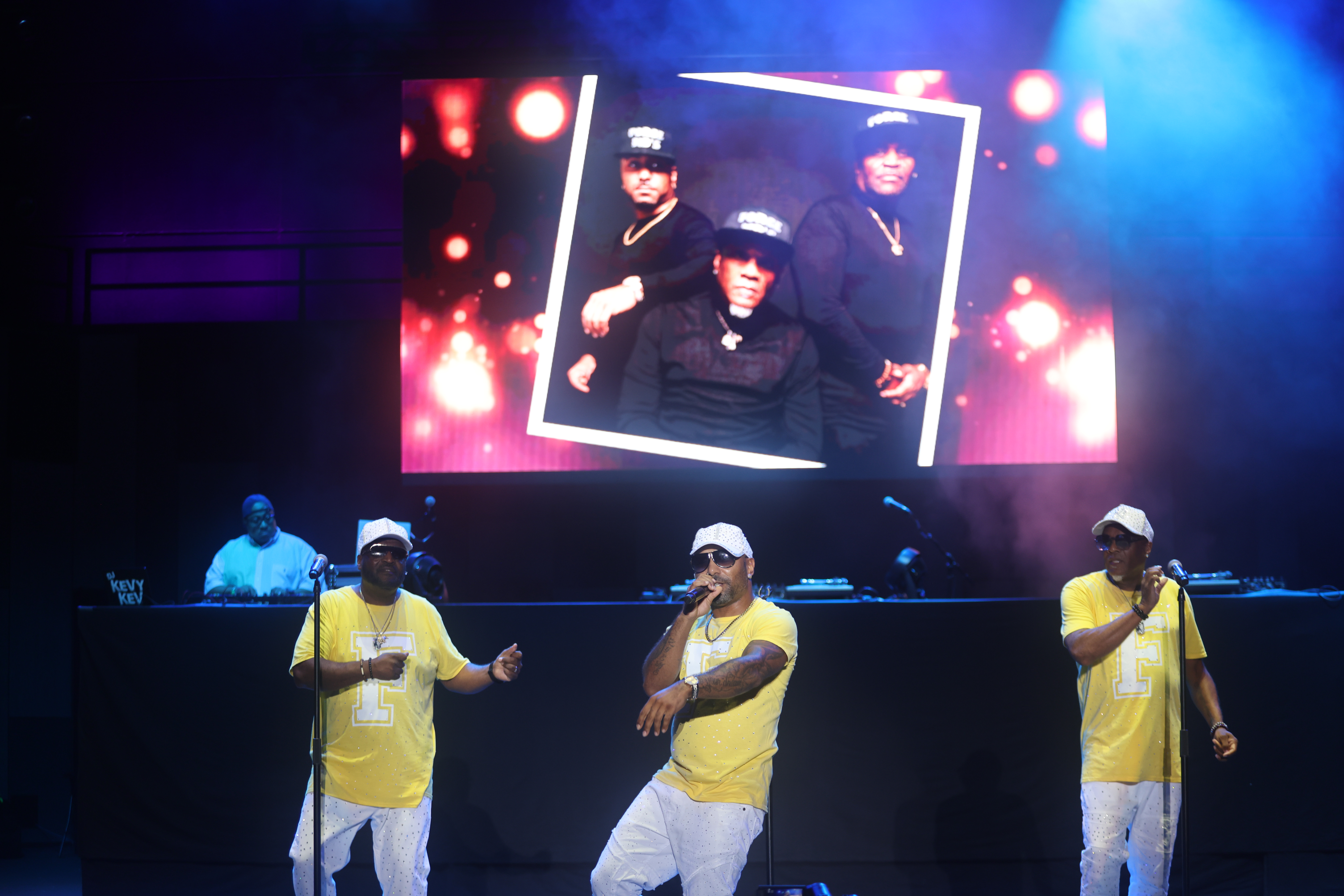 Three men performing on stage, with a large image of the group projected behind them. The men are wearing yellow shirts and appear to be rapping or singing.