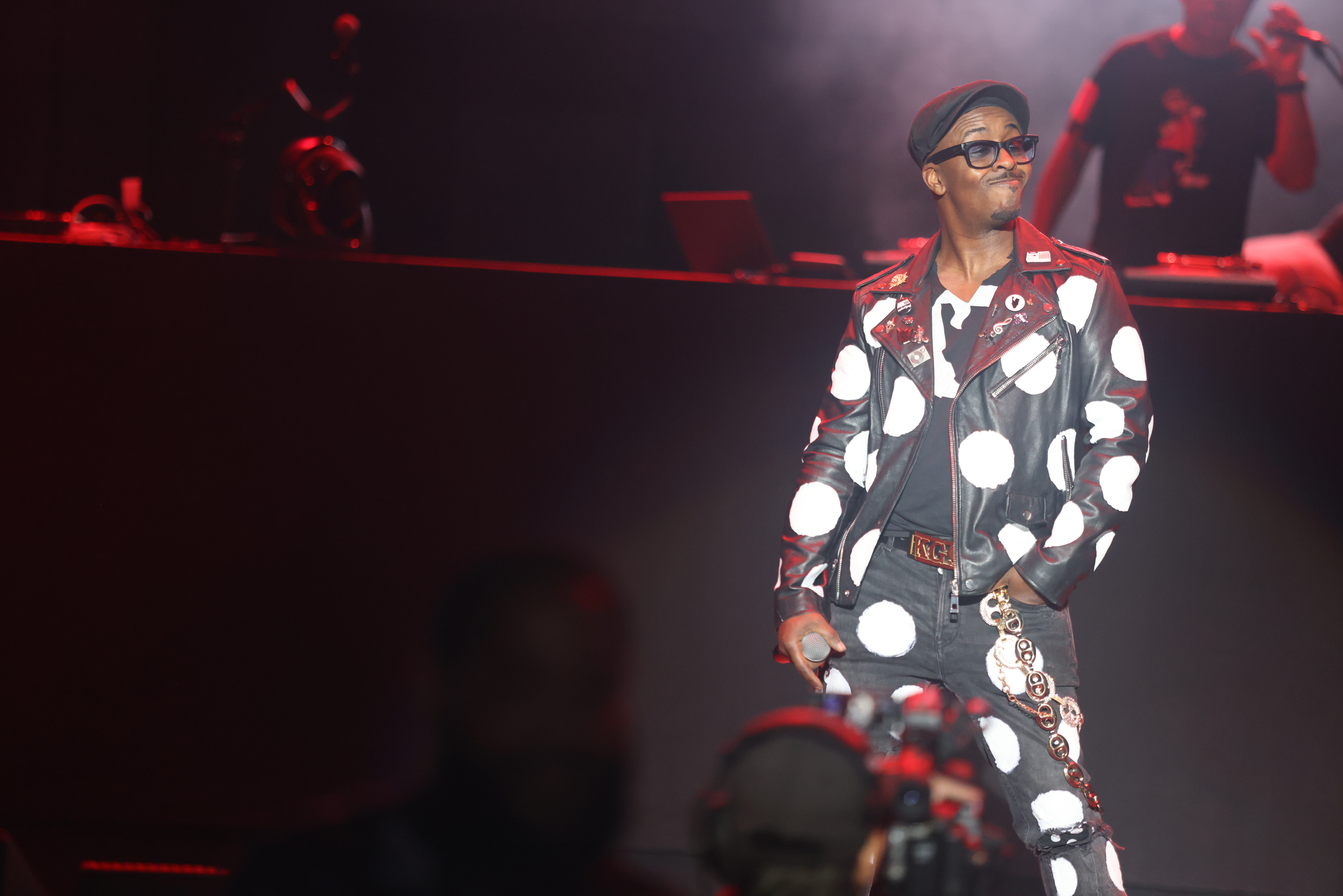 A performer wearing a polka dot jacket and glasses stands on a dimly lit stage, surrounded by smoke and red lighting.