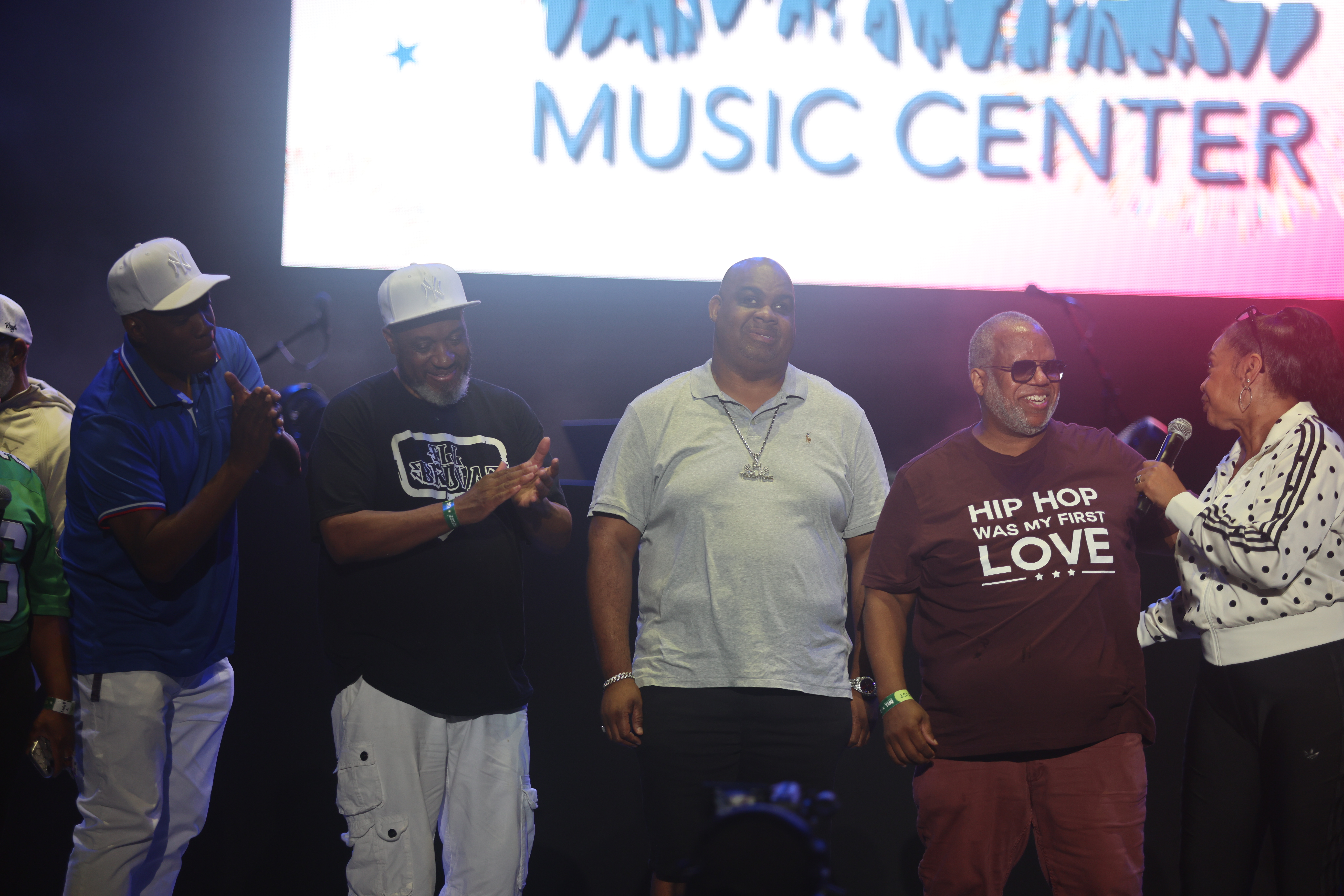 A group of men standing in front of a large "Music Center" sign, some wearing hats and t-shirts with hip hop-related text.