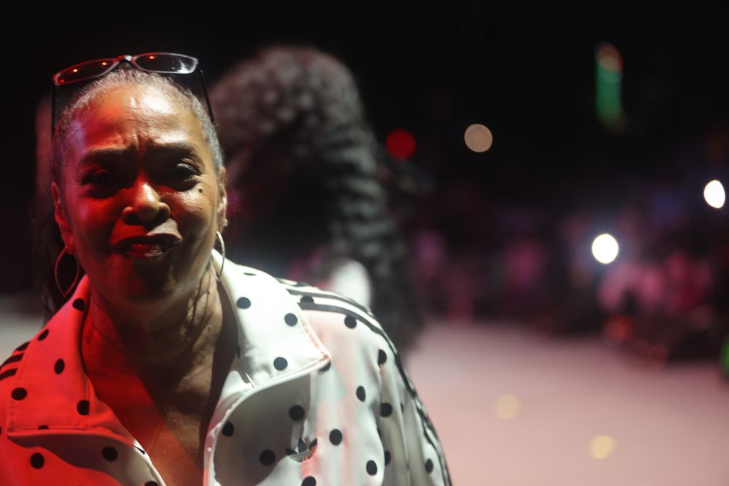 A close-up portrait of an older African American woman wearing a polka dot blouse and red-framed glasses, with a serious expression on her face against a dark background with colorful lights.