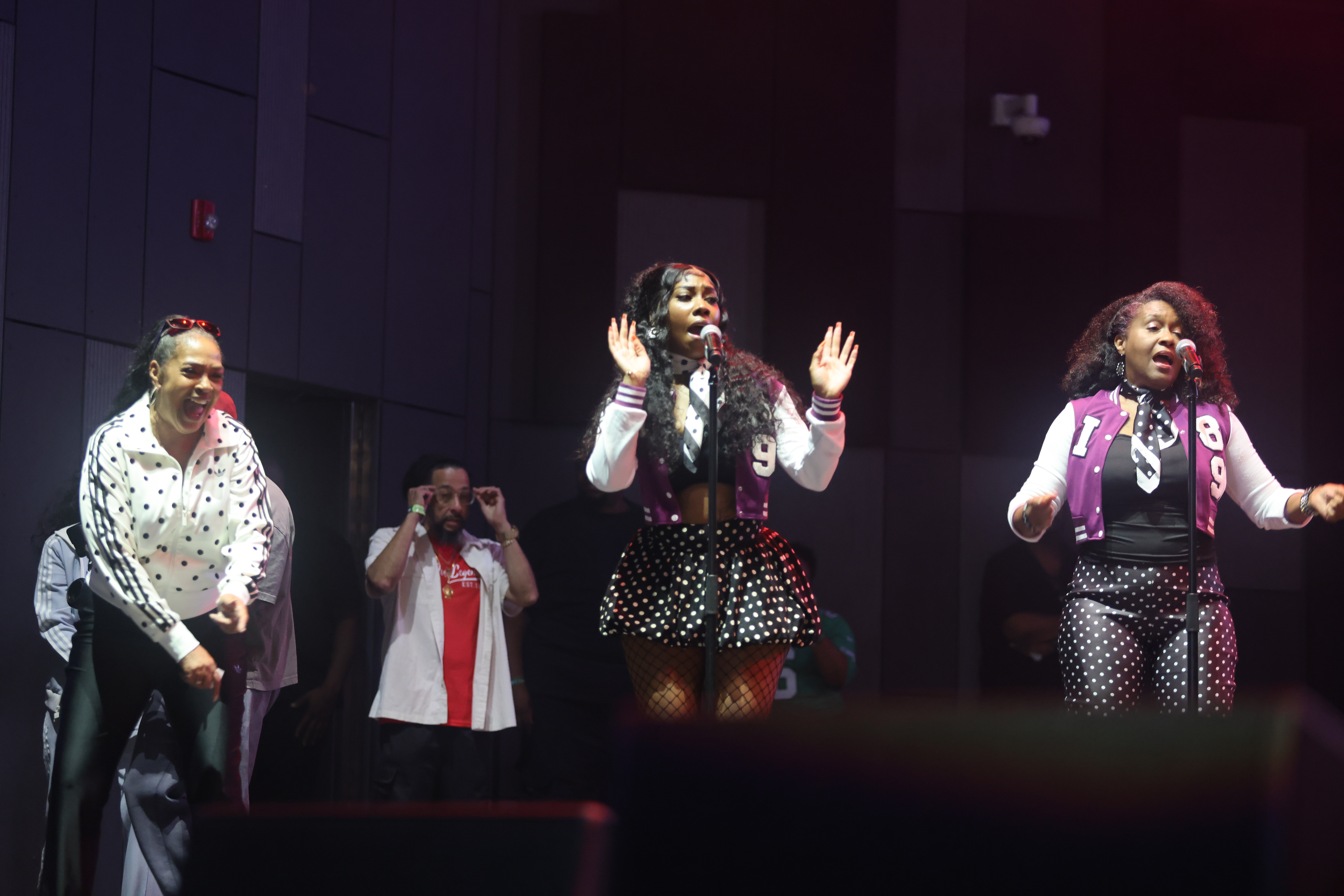 Three performers on stage singing and dancing, wearing polka dot outfits.