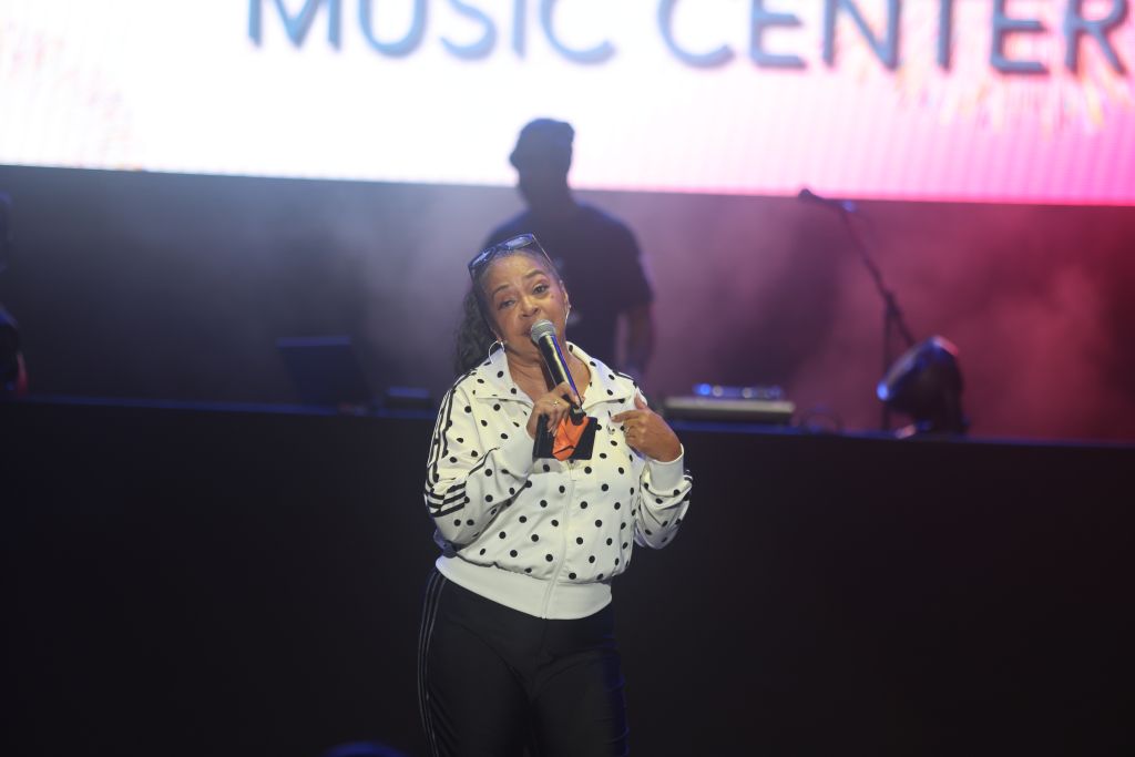 A woman in a polka dot jacket singing into a microphone on a stage with "Music Center" displayed in the background.