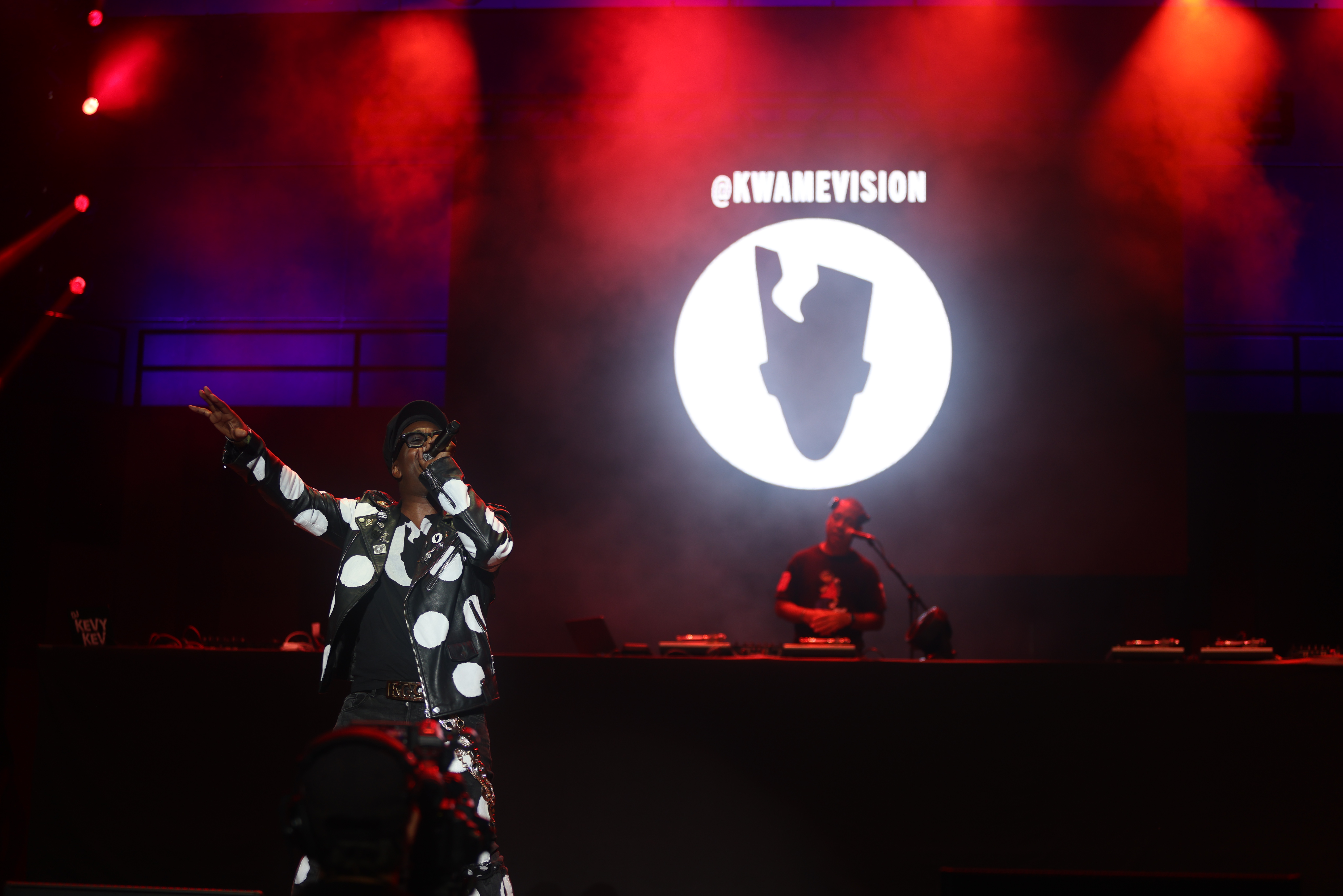 A performer on stage in front of a large projection displaying the text "KWAMEVISION" against a red and purple lighting backdrop.