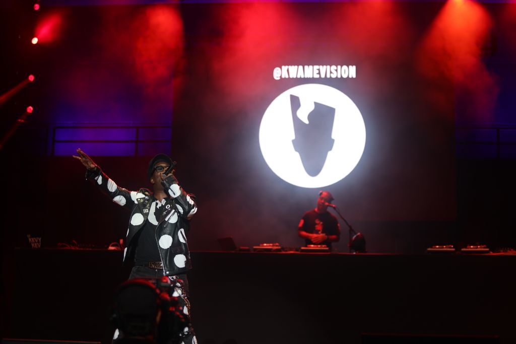 A performer on stage in front of a large projection displaying the text "KWAMEVISION" against a red and purple lighting backdrop.