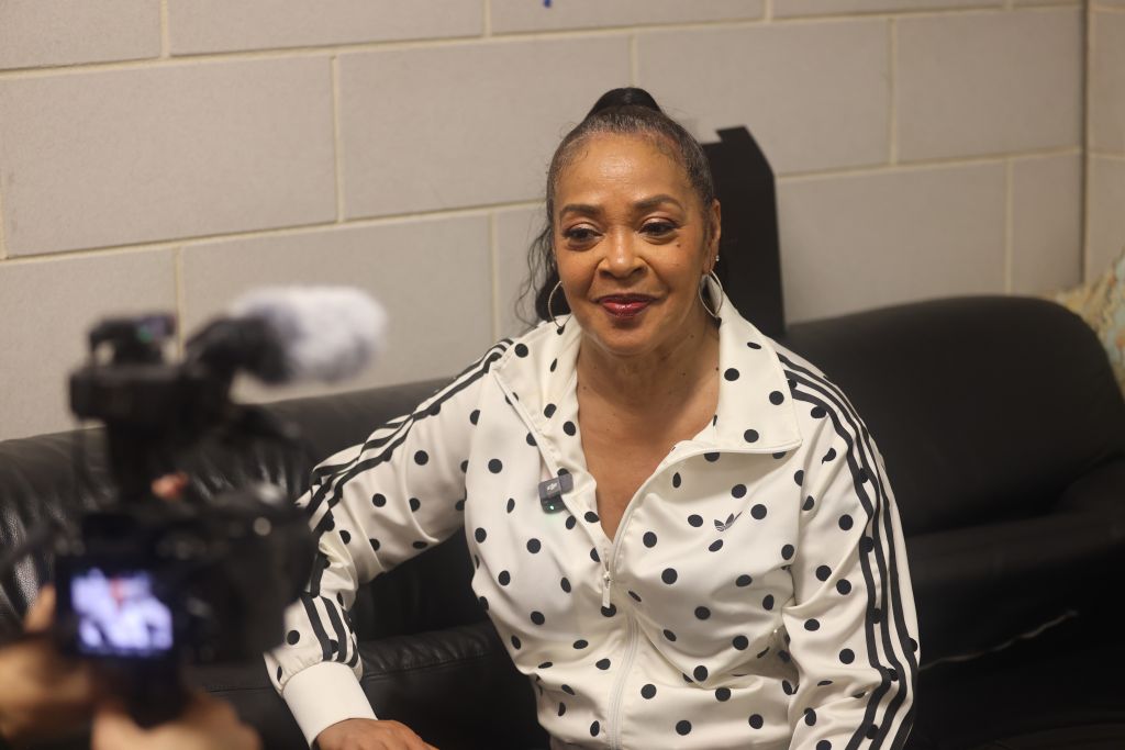 A smiling woman wearing a polka dot and striped blouse sits on a couch.