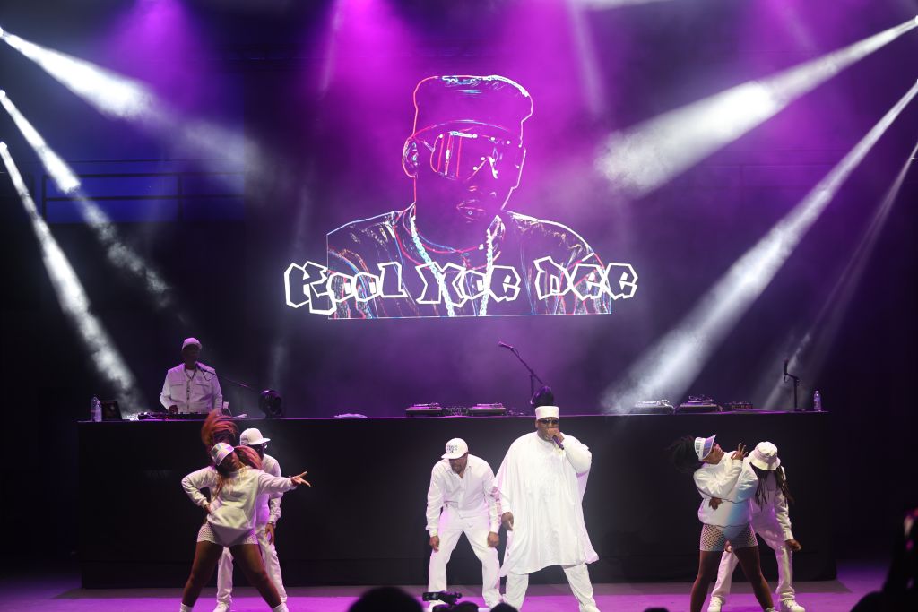 A stage with performers in white outfits dancing, with a large projection of "Coolyrgb" text and a silhouetted figure above them.
