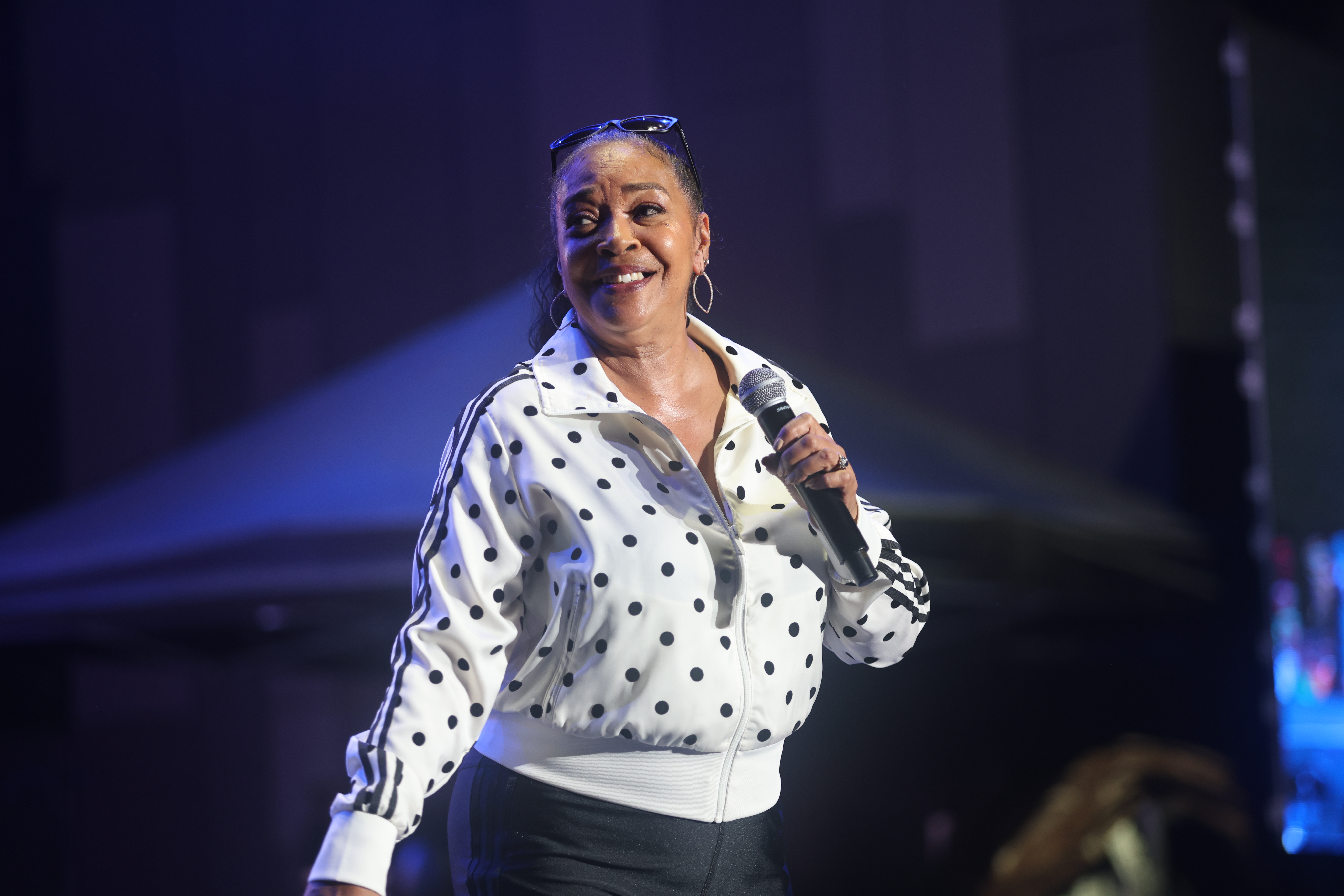 A smiling woman wearing a polka dot shirt and holding a microphone on stage in a dimly lit performance venue.