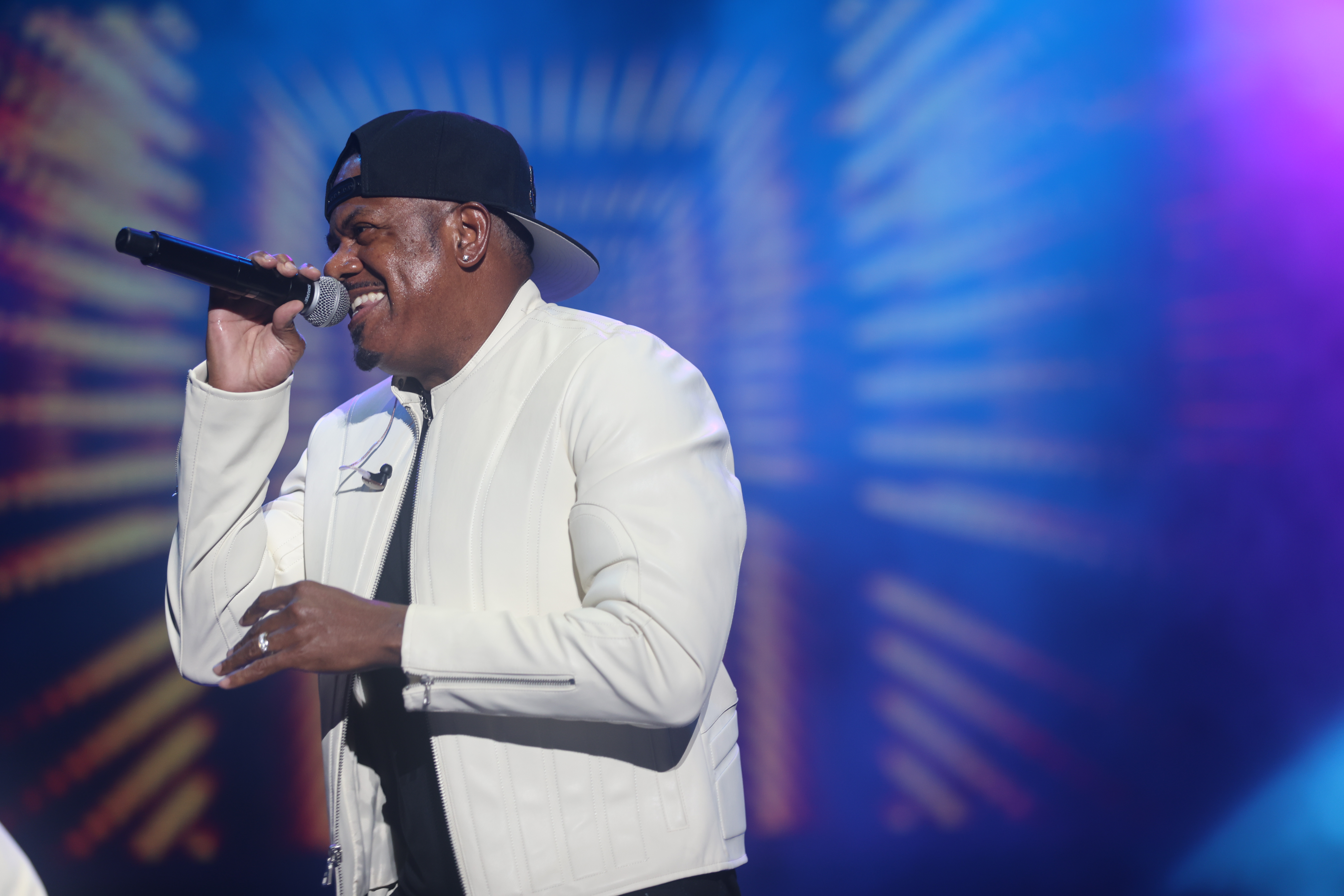 A man wearing a black cap and white suit singing into a microphone on a brightly lit stage.
