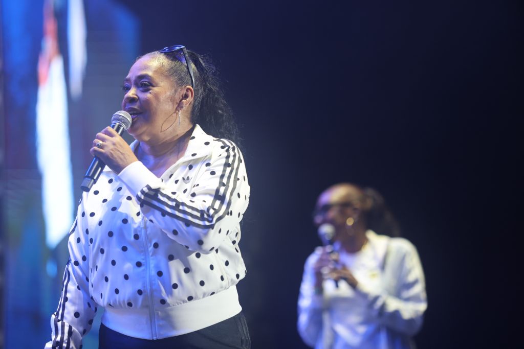 A woman in a polka dot jacket singing into a microphone on stage, with another person in the background.