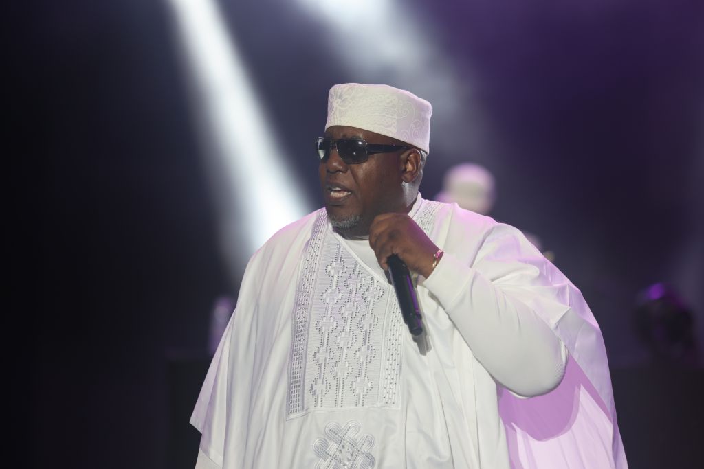 A man wearing a white robe and hat speaking into a microphone on stage, with spotlights shining on him.