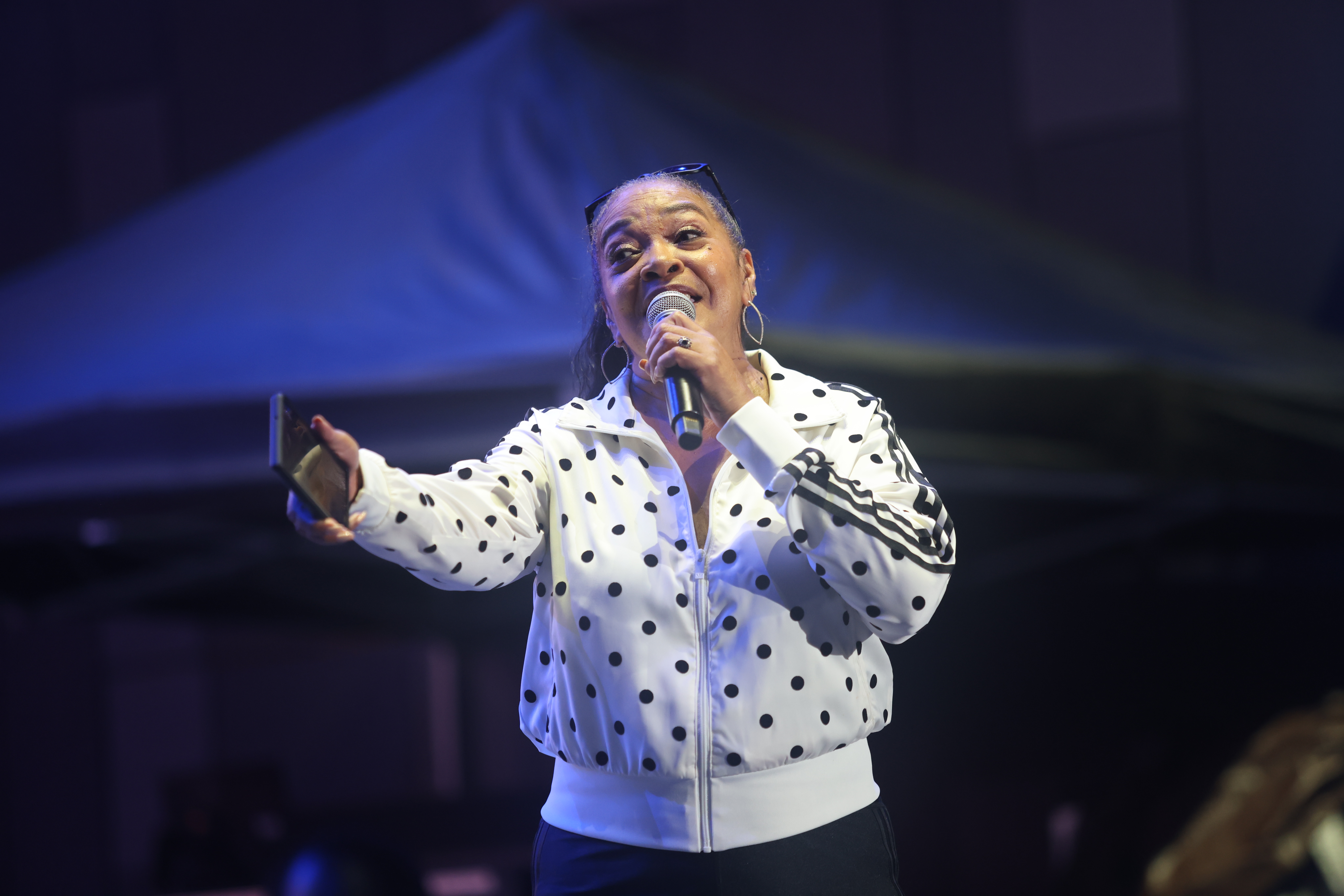 A woman in a polka dot jacket singing into a microphone on stage under blue lighting.