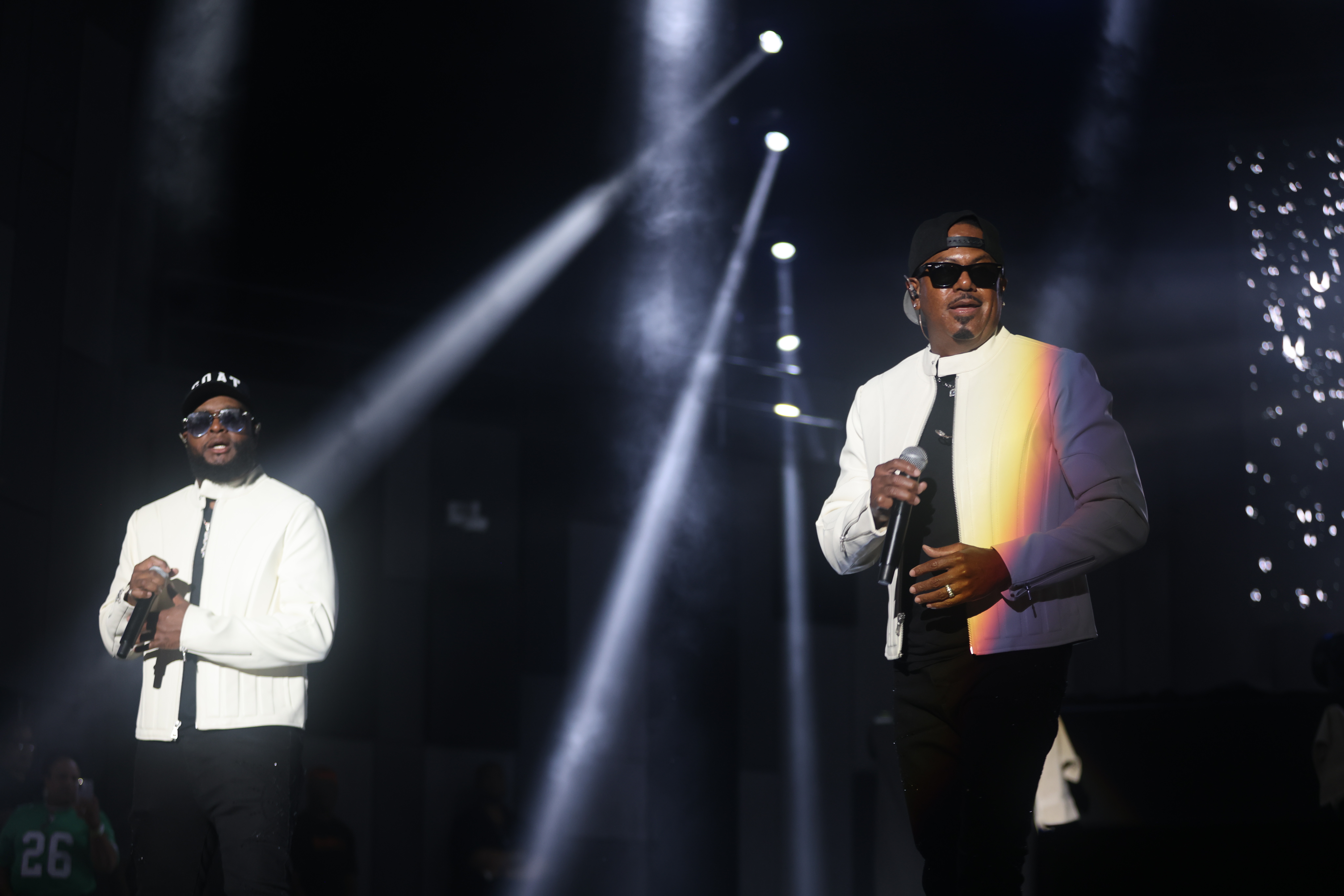 Two performers on stage in white outfits and sunglasses, singing into microphones under bright spotlights.