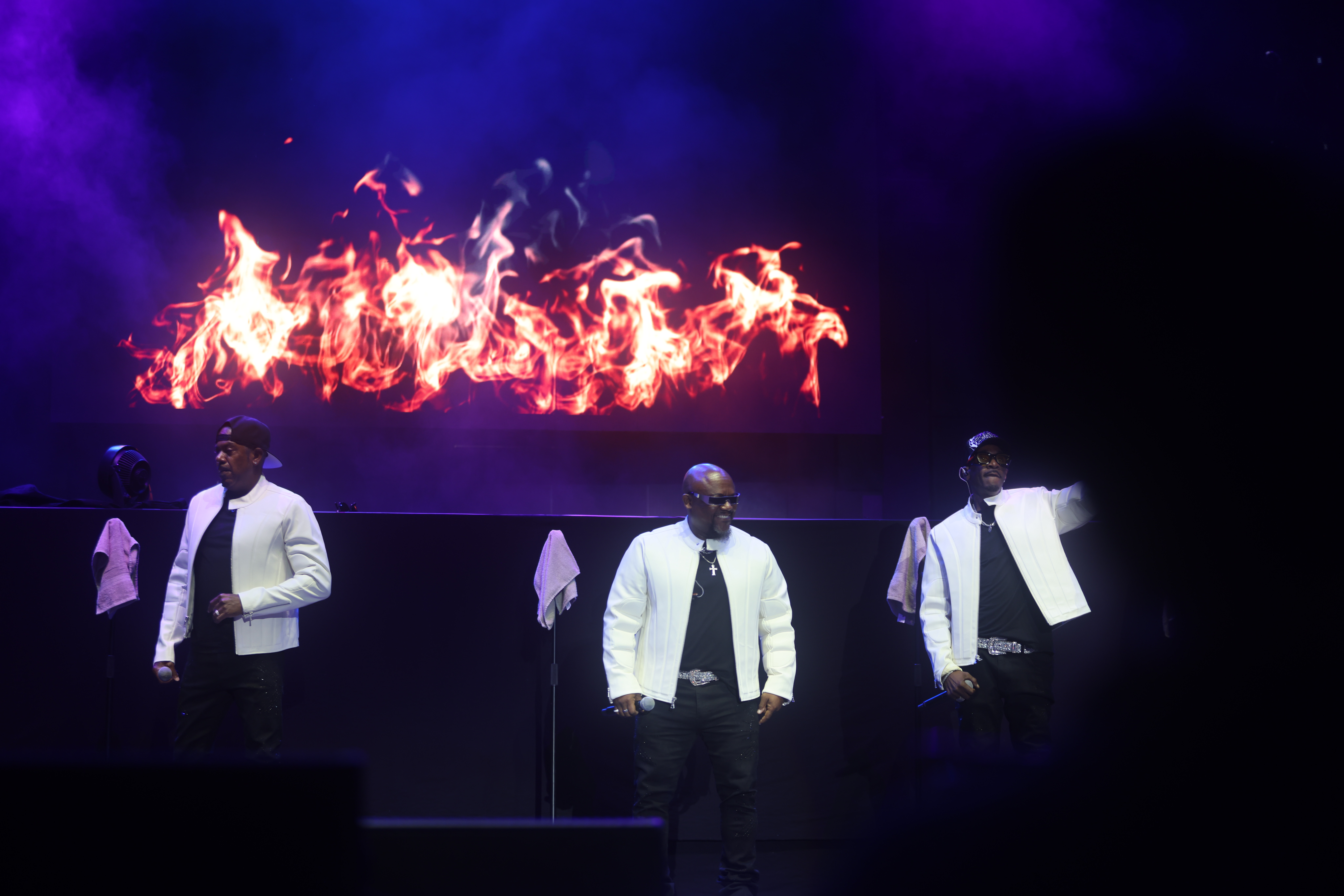 Three performers on stage in white suits, with a large fiery display in the background.