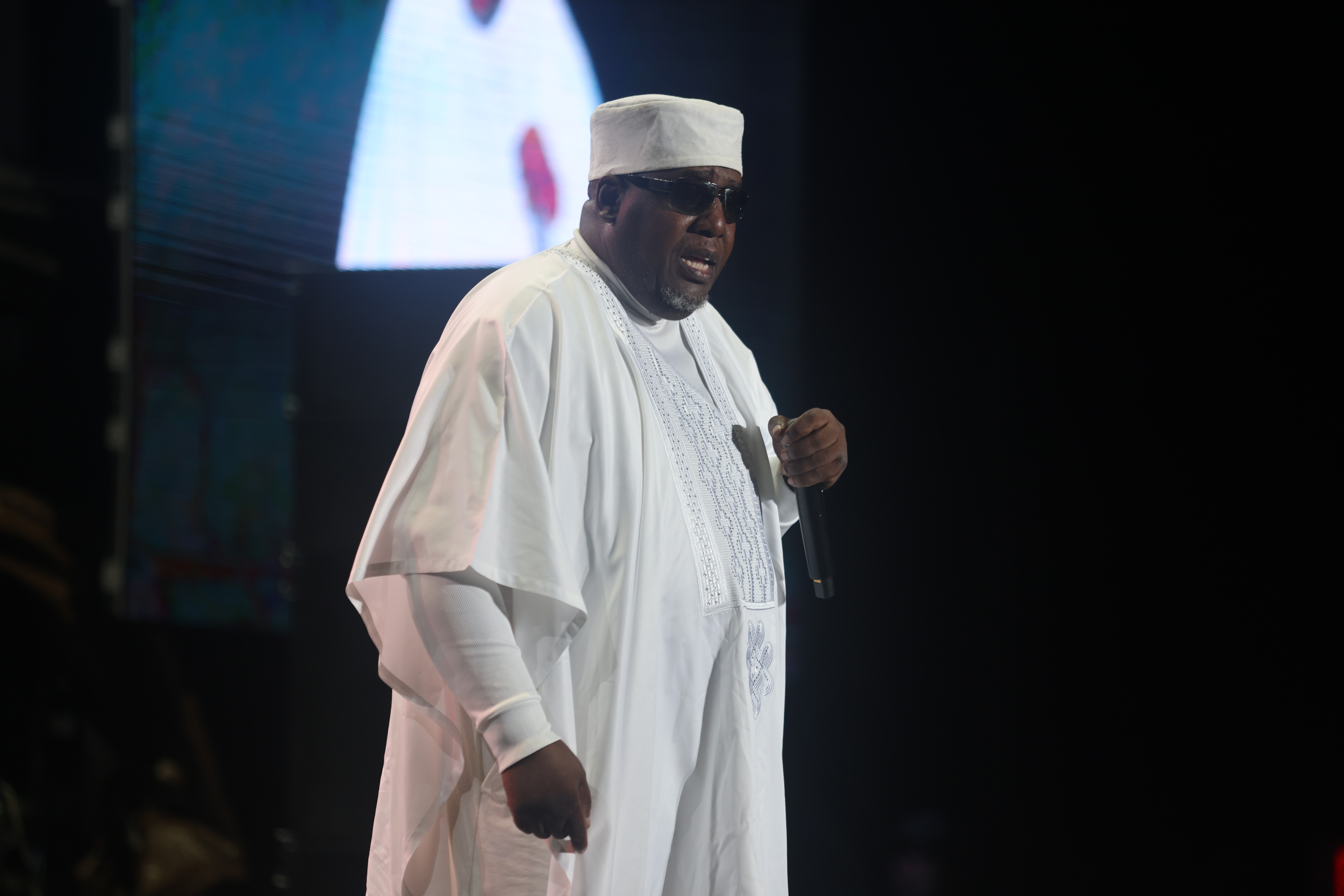 A man wearing a white robe and hat speaking into a microphone on stage.